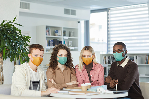 Multi-ethnic group of young people wearing masks and looking at camera while studying together at table in college library, copy space