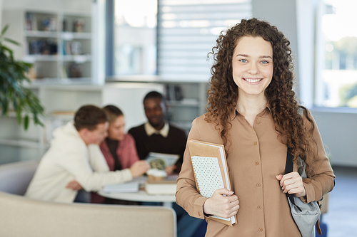 Waist up portrait of curly haired girl looking at camera and smiling while standing in college library with people working in background, copy space