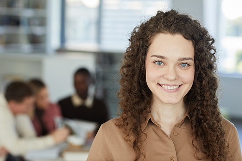 Close up portrait of curly haired girl looking at camera and smiling while standing in college library with people working in background, copy space