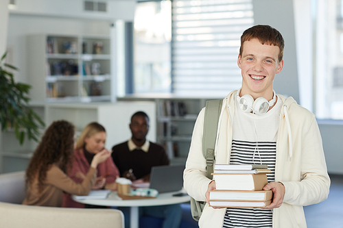 Waist up portrait of smiling teenage boy looking at camera while standing in college library with people working in background, copy space