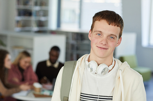 Head and shoulders portrait of smiling teenage boy looking at camera while standing in college library with people working in background, copy space