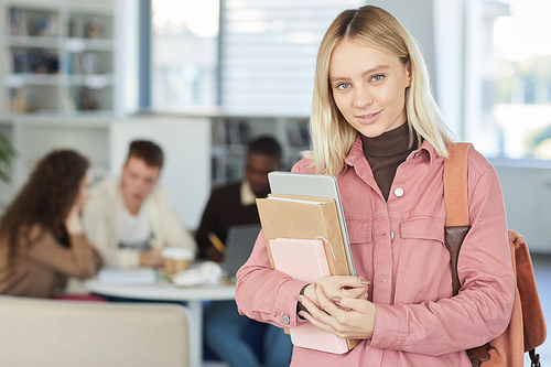 Waist up portrait of blonde young woman looking at camera and smiling while standing in college library with people working in background, copy space