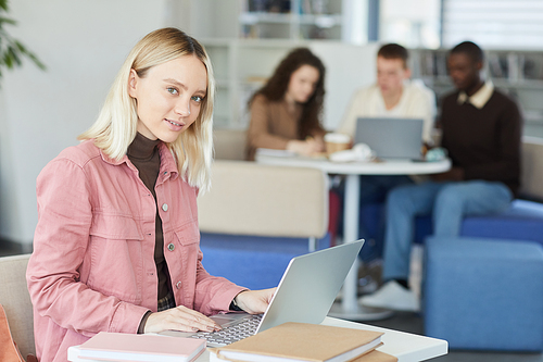 Side view portrait of blonde young woman looking at camera while using laptop in college library with people in background, copy space