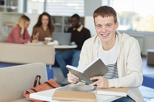 Portrait of smiling red haired boy looking at camera while studying in college library and holding textbook, copy space