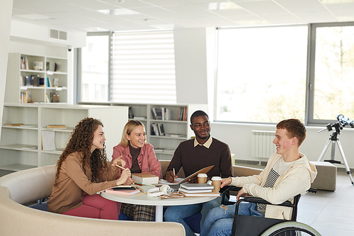 Side view at multi-ethnic group of students studying in college library featuring young man using wheelchair in foreground, copy space