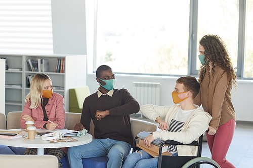 Side view at multi-ethnic group of students wearing masks while studying in college library with young man using wheelchair in foreground, copy space