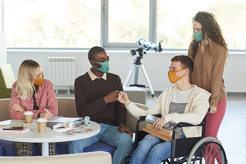 Portrait of multi-ethnic group of students wearing masks while studying in college library with young man using wheelchair in foreground, copy space