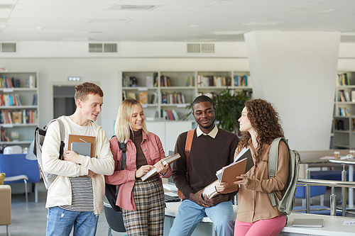 Portrait of multi-ethnic group of students standing in college library and chatting while holding books and backpacks, copy space above