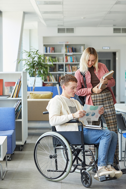 Vertical full length portrait of cheerful disabled student talking to young woman in college library