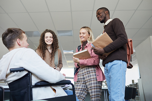 Low angle view at multi-ethnic group of students talking to young man in wheelchair while studying together in college library