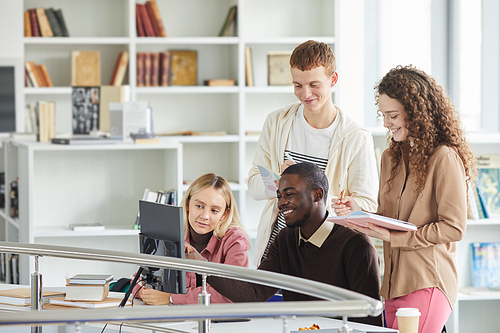 Multi-ethnic group of students using telecommunication equipment while studying in college library and smiling, copy space