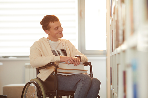 Warm toned portrait of young man using wheelchair in school while looking at bookshelves in library lit by sunlight