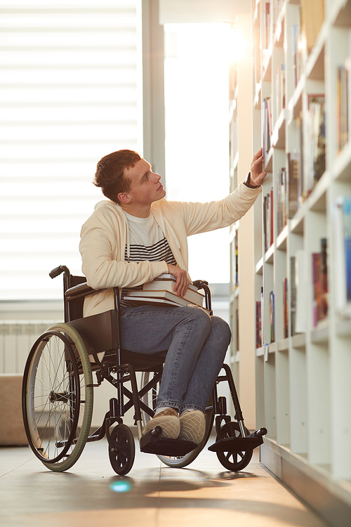 Vertical full length portrait of young man using wheelchair in school while looking at bookshelves in library lit by sunlight