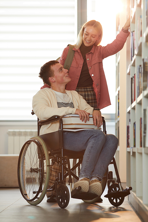 Vertical full length portrait of young man using wheelchair in school with female friend helping him in library lit by sunlight