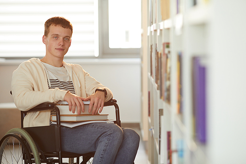 Warm toned portrait of young man using wheelchair in school while looking at camera in library lit by sunlight