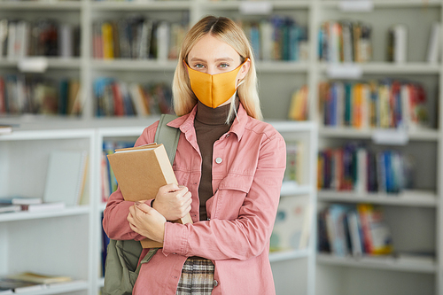 Waist up portrait of blonde young woman wearing mask while standing in school library and holding books, copy space