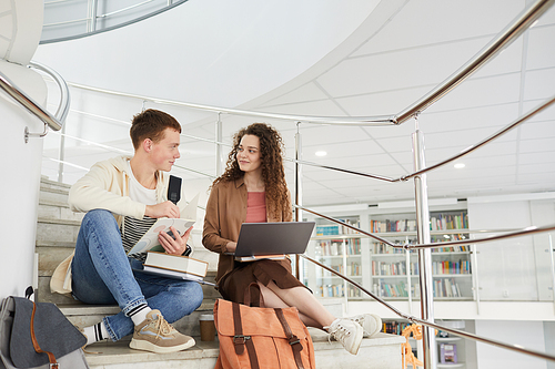 Wide angle view at two students sitting on stairs in college library and using laptop while working on homework, copy space