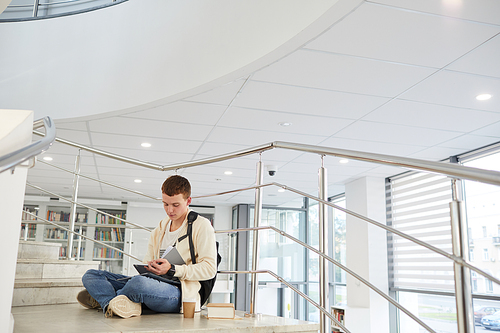 Wide angle view at young man sitting on stairs in college library and working on homework, copy space