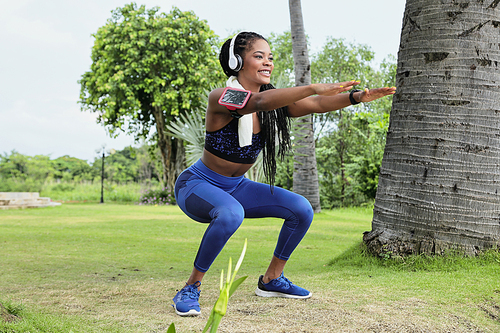 Happy beautiful young Black sportwoman in headphones doing squats in park after morning run