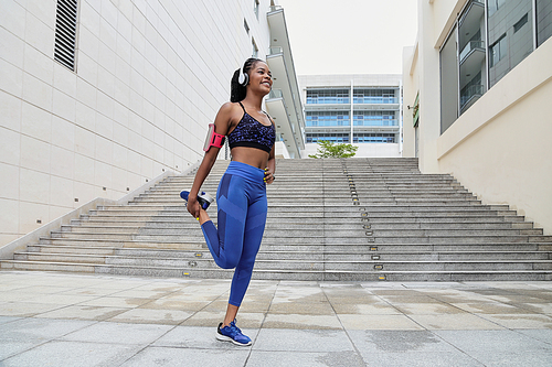 Beautiful happy African-American sportswoman in headphones standing on one leg and stretching quadriceps