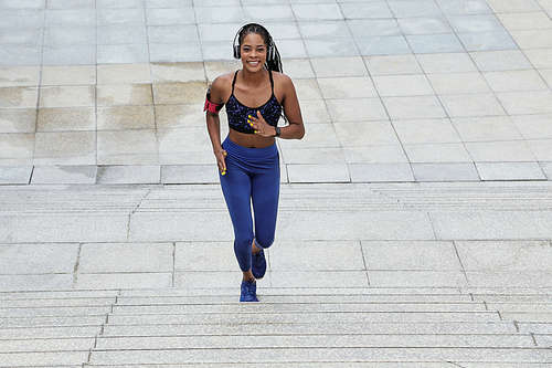 Happy pretty Black female jogger in top and leggins running up the stairs and looking at camera