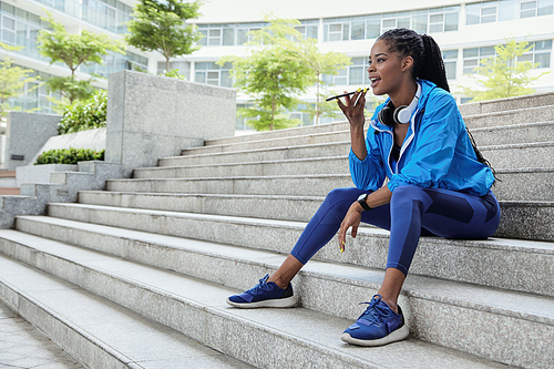 Pretty fit mixed-race young sportswoman sitting on steps and recording voice message for her friend or trainer