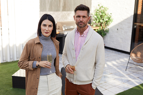 Waist up portrait of contemporary adult couple looking at camera while standing at outdoor terrace holding champagne flutes, copy space