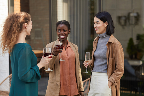 Multi-ethnic group of contemporary adult women chatting while enjoying wine at outdoor party, copy space