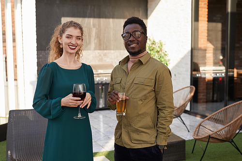 Waist up portrait of contemporary mixed-race couple looking at camera while enjoying wine standing at outdoor terrace, copy space