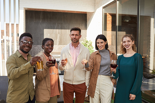 Waist up portrait of multi-ethnic group of friends looking at camera while enjoying wine standing at outdoor terrace during party