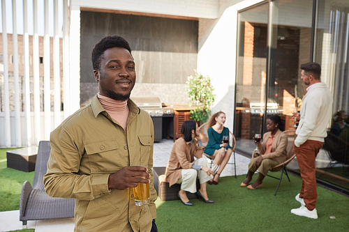 Waist up portrait of young African-American man looking at camera and smiling while standing at terrace during outdoor party, copy space