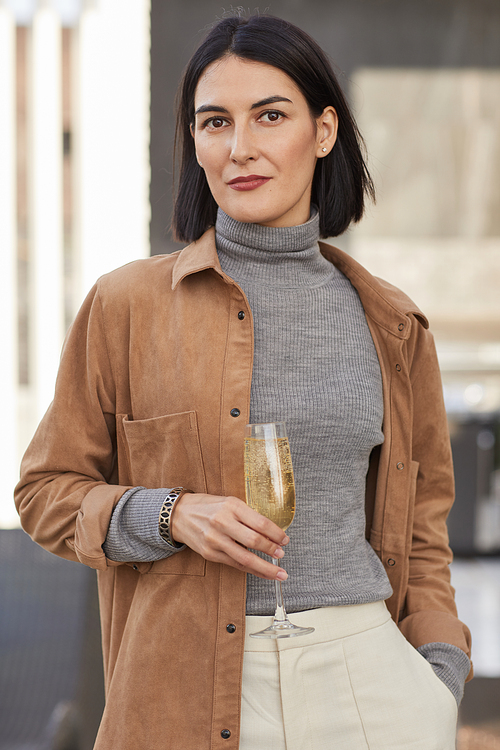 Vertical waist up portrait of elegant contemporary woman holding champagne flute and looking at camera while enjoying party outdoors