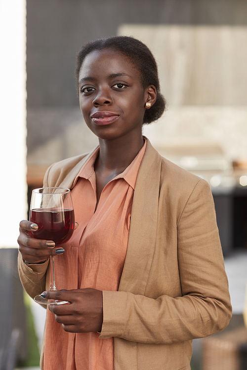 Vertical waist up portrait of elegant African-American woman holding wine glass and looking at camera while enjoying party outdoors