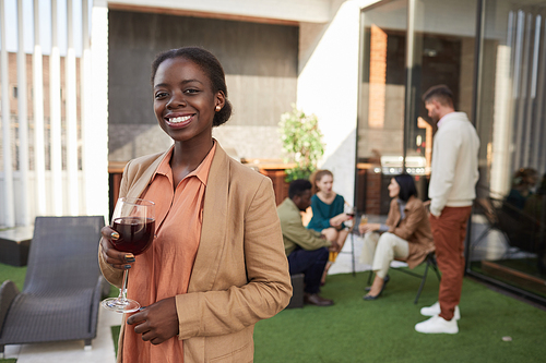 Waist up portrait of elegant African-American woman holding wine glass and looking at camera while enjoying party outdoors, copy space