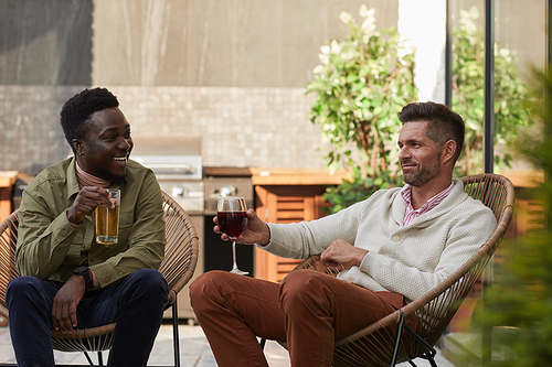 Portrait of two contemporary men enjoying wine while relaxing in lounge chairs at outdoor terrace, copy space