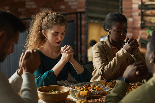 Warm toned portrait of multi-ethnic group of elegant young people praying with eyes closed while sitting at dinner table during Thanksgiving celebration, copy space