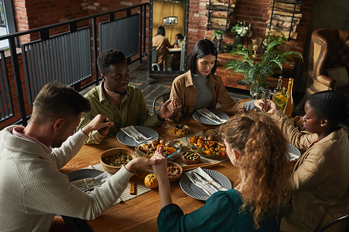 High angle portrait of multi-ethnic group of elegant young people praying and holding hands while sitting at dinner table during Thanksgiving celebration, copy space