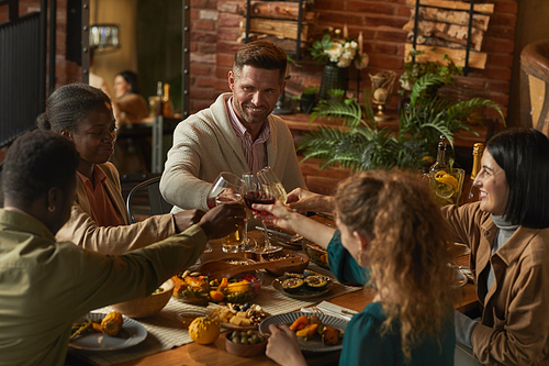 Multi-ethnic group of people clinking glasses while enjoying dinner party with friends and family in cozy interior