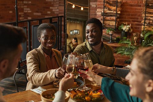 Portrait of young African-American couple clinking glasses while enjoying dinner party with friends and family in cozy interior