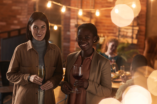 Waist up portrait of two young women holding champagne while enjoying outdoor party with friends