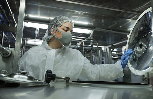 Portrait of young female worker wearing mask and protective wear opening barrels while working at food factory, copy space