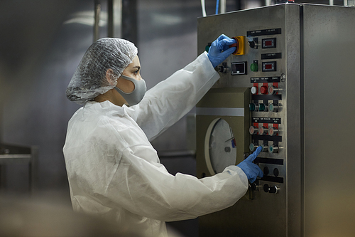 Side view portrait of young woman pushing buttons on control panel while operating machine units at food factory, copy space