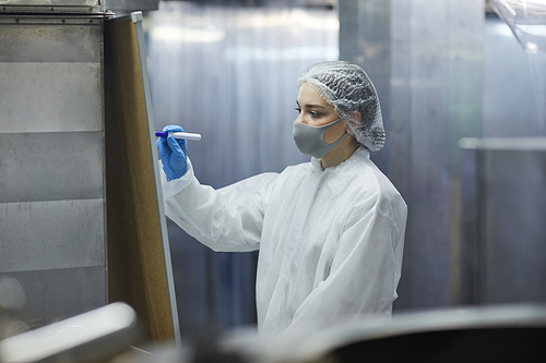 Portrait of female worker wearing mask and protective suit while writing on board in factory workshop, copy space