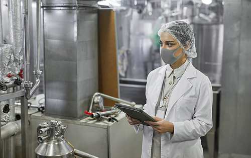 Waist up portrait of female worker wearing mask and holding digital tablet during quality control inspection at food factory, copy space