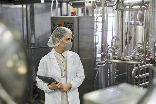 Side view portrait of female worker wearing mask and holding digital tablet during quality control inspection at food factory, copy space