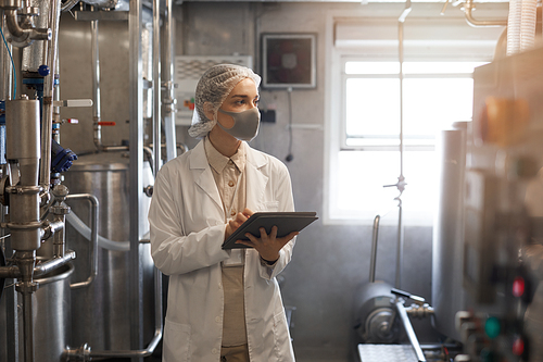 Waist up portrait of young woman wearing mask and holding digital tablet during quality control inspection in sunlit workshop at food factory, copy space