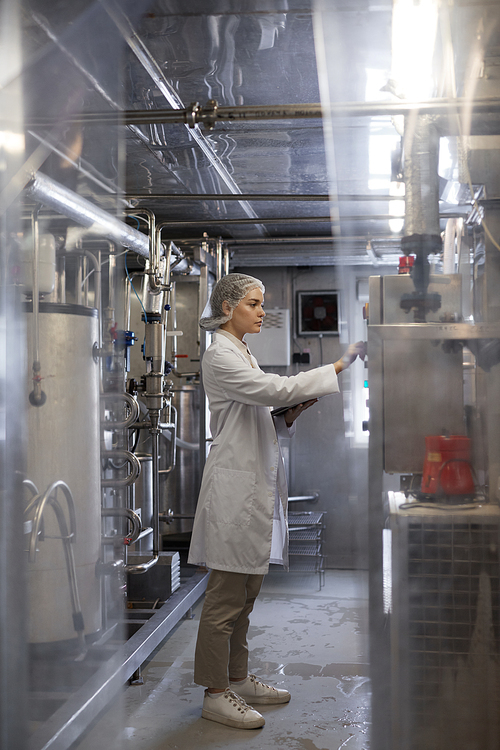 Vertical full length portrait of young woman operating machine units while working at industrial food factory