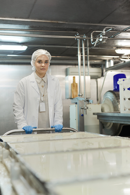 Vertical portrait of young female worker pushing cart with raw cheese in workshop at food factory and looking at camera, copy space