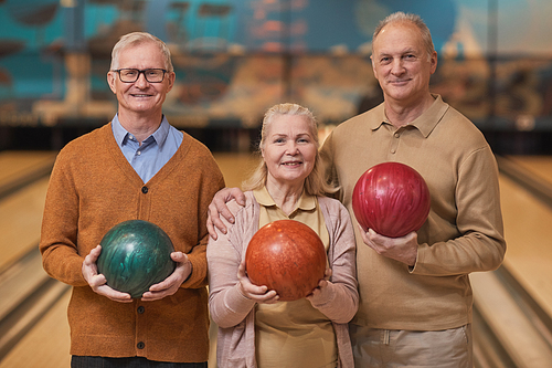 Waist up portrait of three smiling senior people holding bowling balls and looking at camera while enjoying active entertainment at bowling alley, copy space