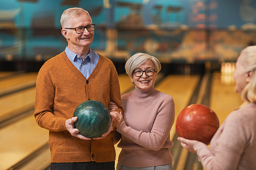 Waist up portrait of group of happy senior people holding bowling balls and chatting while enjoying active entertainment at bowling alley, copy space